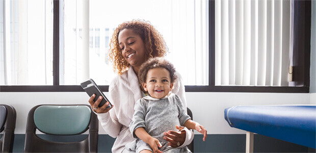 Smiling mother holding her young child while looking at a smartphone in a healthcare setting, with a patient chair and examination table in the background.
